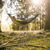 A person relaxes in a large hammock with a dog in a sunlit forest clearing, with trees, moss, and branches visible around them.