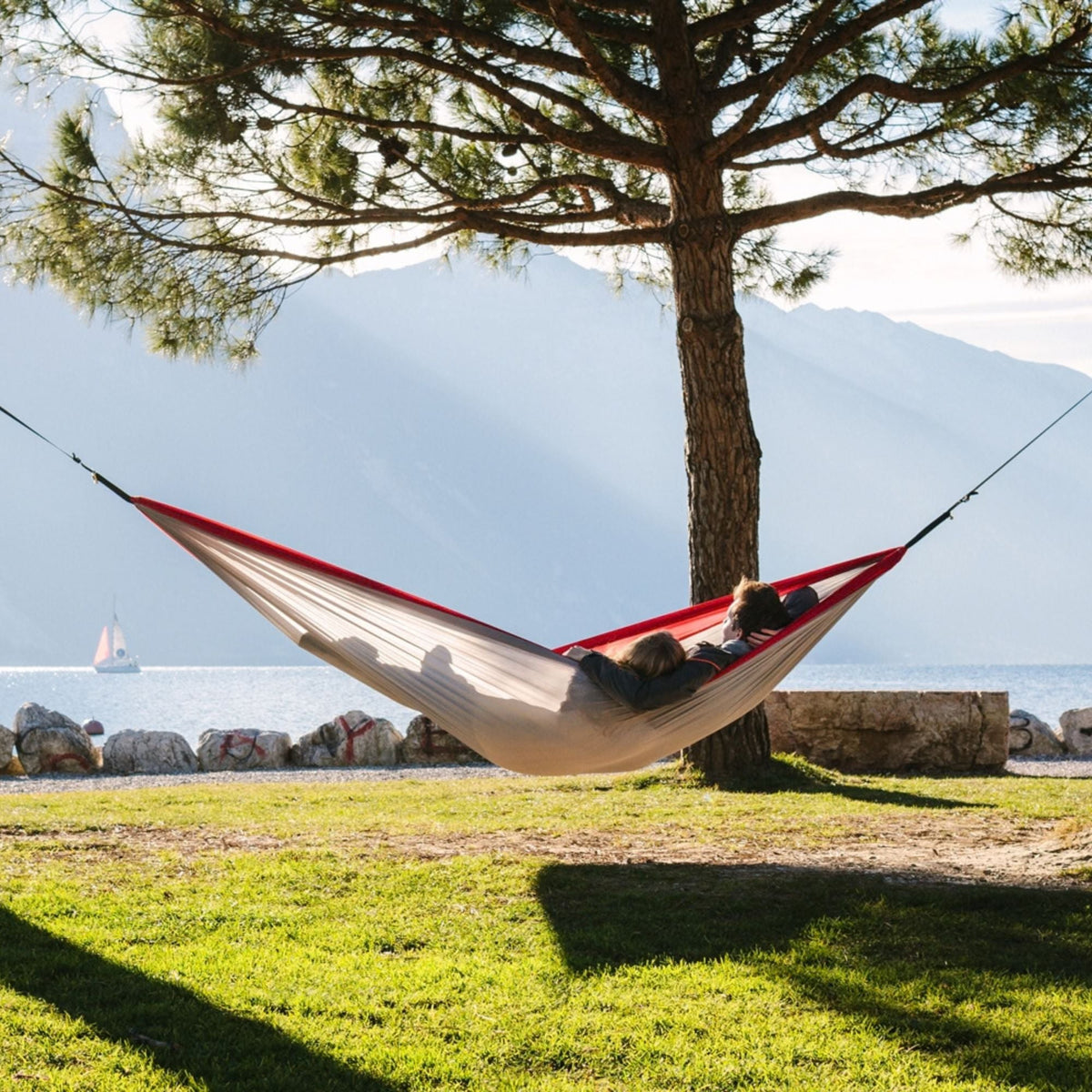 A red and grey hammock is suspended between two trees in a grassy outdoor area near a lake, with mountains in the background. Two people are relaxing in the hammock.
