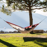 A red and grey hammock is suspended between two trees in a grassy outdoor area near a lake, with mountains in the background. Two people are relaxing in the hammock.