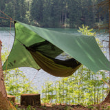 A green camping tarp suspended between trees above a hammock, set up in a forest campsite next to a lake with trees in the background.