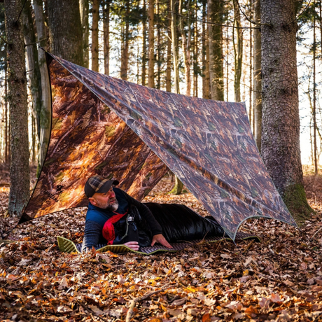 A man lies on the ground under a camouflage tarp shelter in a forest of tall trees, surrounded by fallen autumn leaves.