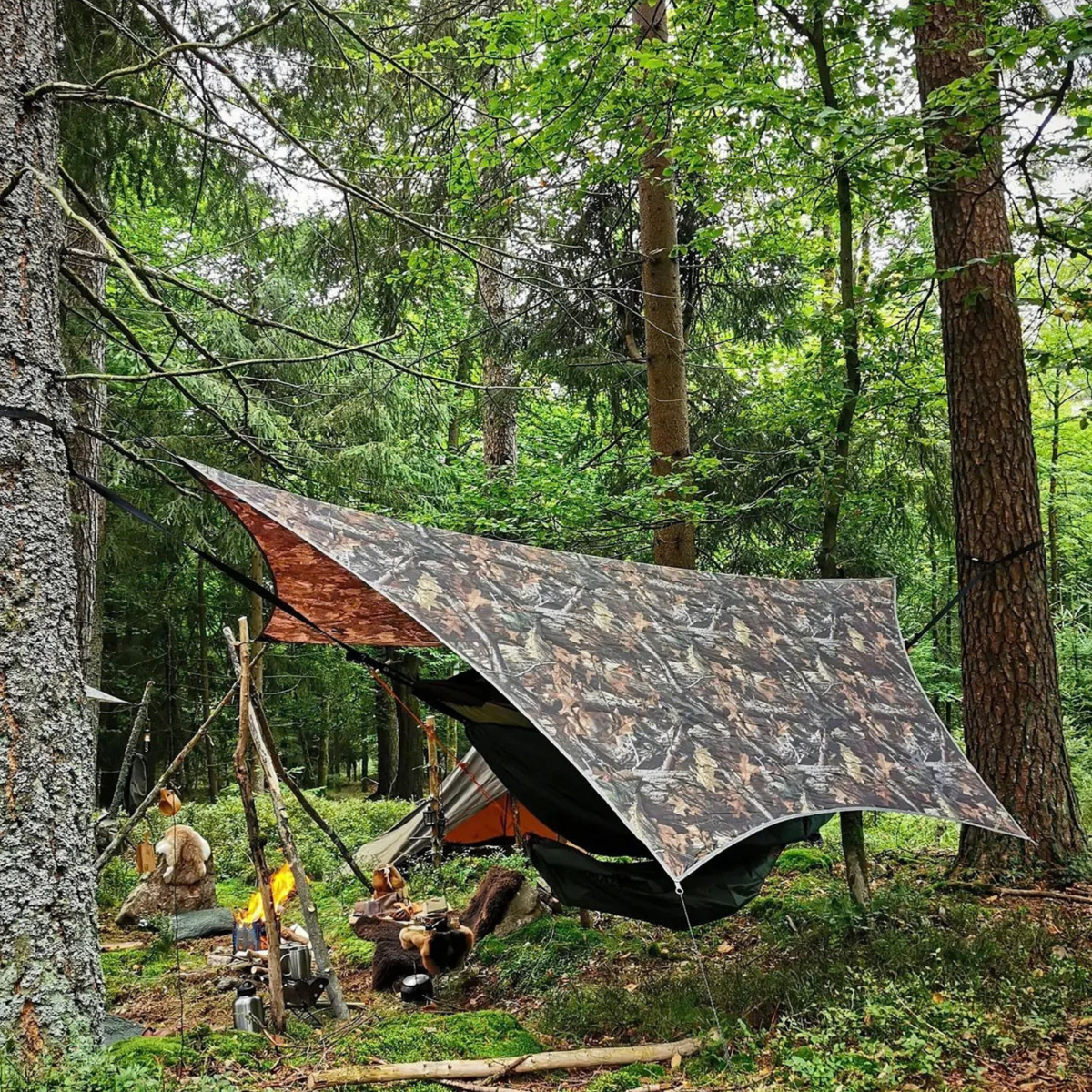 A camouflage tarp shelter set up in a forest with trees, ropes, a hammock, and a campfire visible below.