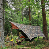 A camouflage tarp shelter set up in a forest with trees, ropes, a hammock, and a campfire visible below.