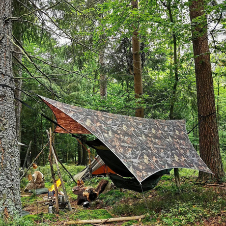A camouflage tarp shelter set up in a forest with trees, ropes, a hammock, and a campfire visible below.