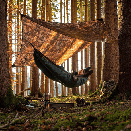 A person relaxes in a green camping hammock suspended between two trees under a large camouflage tarp in a sunlit forest.