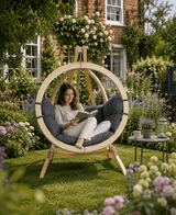 Woman reading a book in a circular wooden chair surrounded by flowers and greenery.