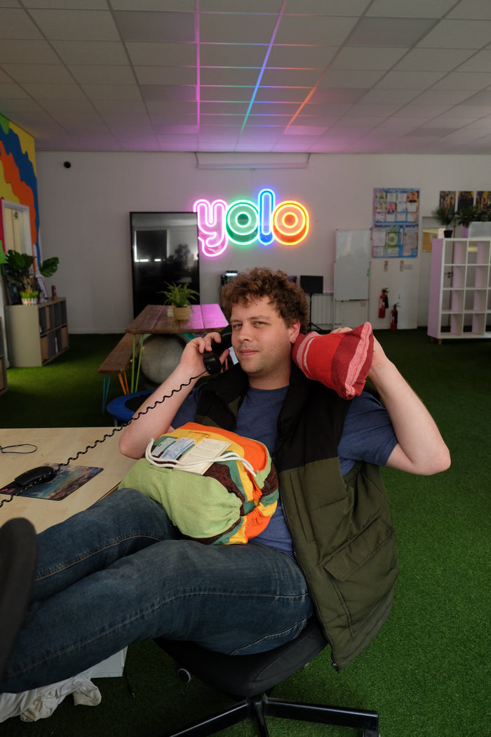 Person sitting on a chair with colourful pillows and a phone, in a room with a 'YOLO' neon sign.