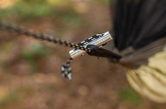 Close-up of a hammock's rope secured with a metallic clip against a blurred forest background. The scene conveys a sense of outdoor relaxation.