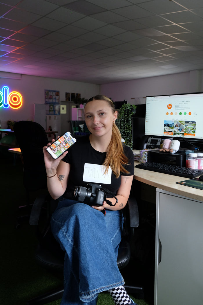 Person holding a camera in an office setting with a computer monitor and desk in the background.
