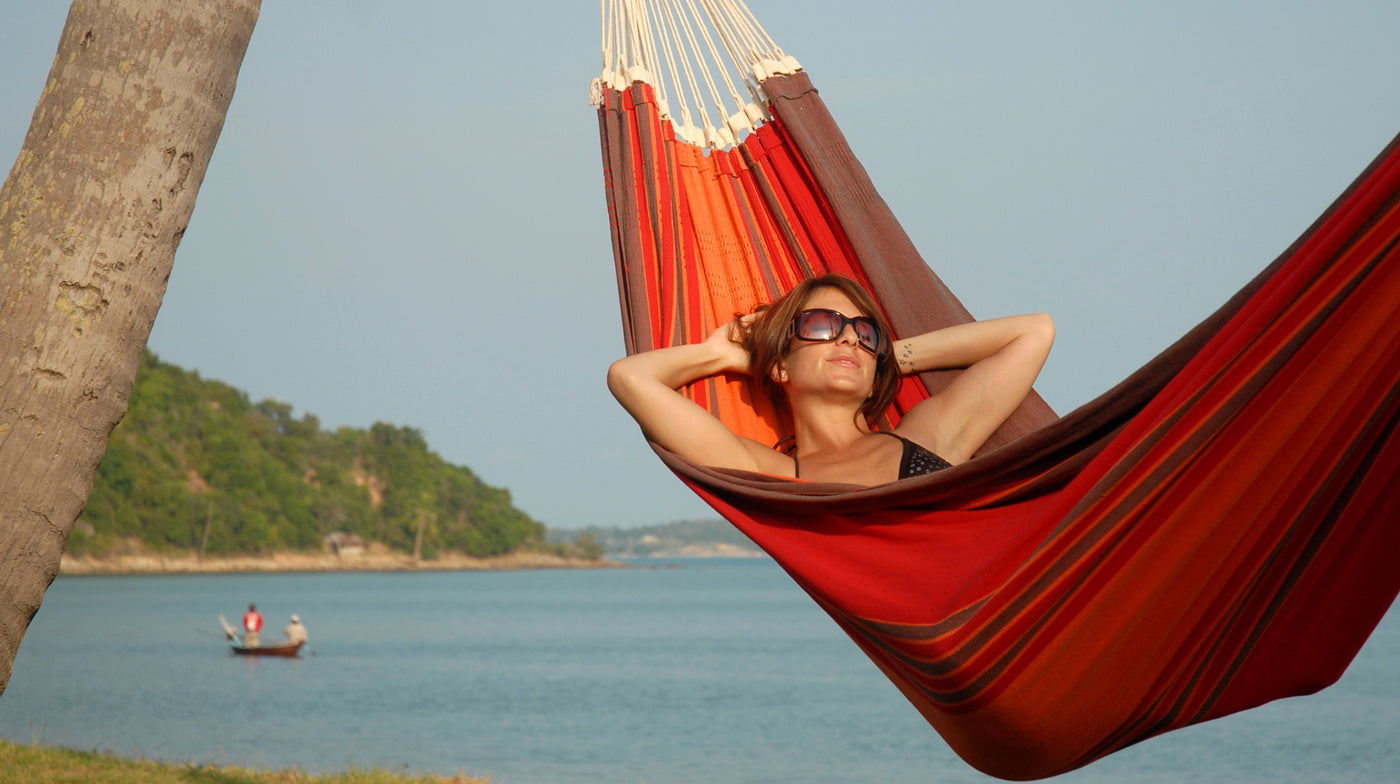 A woman wearing sunglasses relaxes in a red and brown striped hammock strung between trees by a lake, with a small boat and green hills in the background.