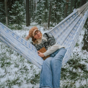 Woman in a blue Carioca hammock sat in a scenic winter forest. 