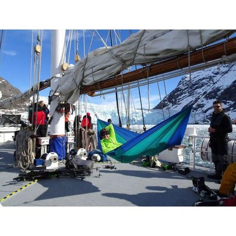 People on a boat with a sail, one person in a green hammock, mountains in the background