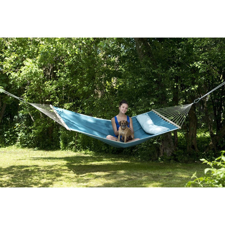 A person sitting in a blue hammock with a dog, surrounded by greenery