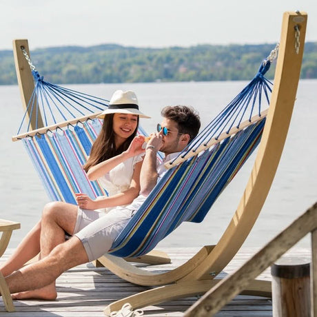 Two people relaxing in a striped hammock by a lake.