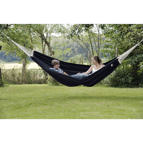 Two people relaxing in a black hammock surrounded by greenery.