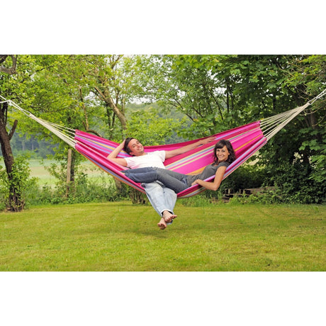 Two people relaxing in a pink and white hammock surrounded by greenery.