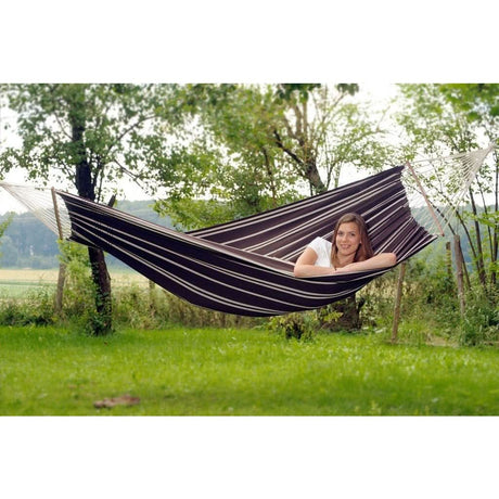 A woman relaxing in a striped hammock in a natural setting with trees and grass.