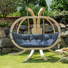 Wooden hanging chair with blue cushion on a grassy area with a stone wall and garden in the background.