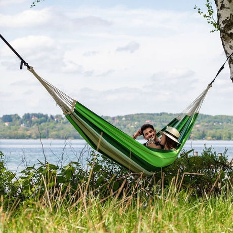 Two people in a green hammock by a lake with trees in the background