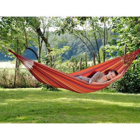 A person relaxing in a red and orange striped hammock in a lush, green outdoor setting.