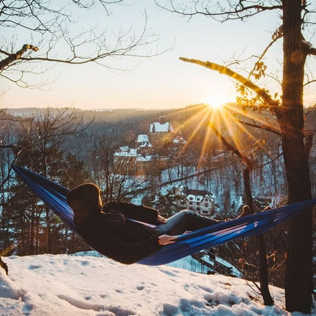 A person in a blue hammock on a snowy landscape with a scenic view during sunset.
