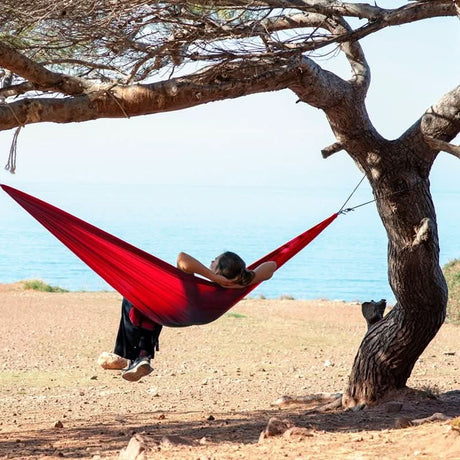 Person relaxing in a red hammock between two trees with a clear sky and ocean in the background.